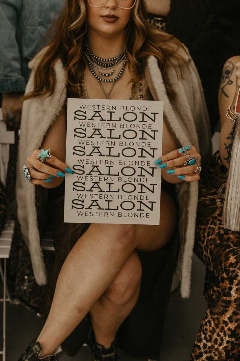 Woman holding "Western Blonde Salon" sign, showcasing bold style and layered jewelry.