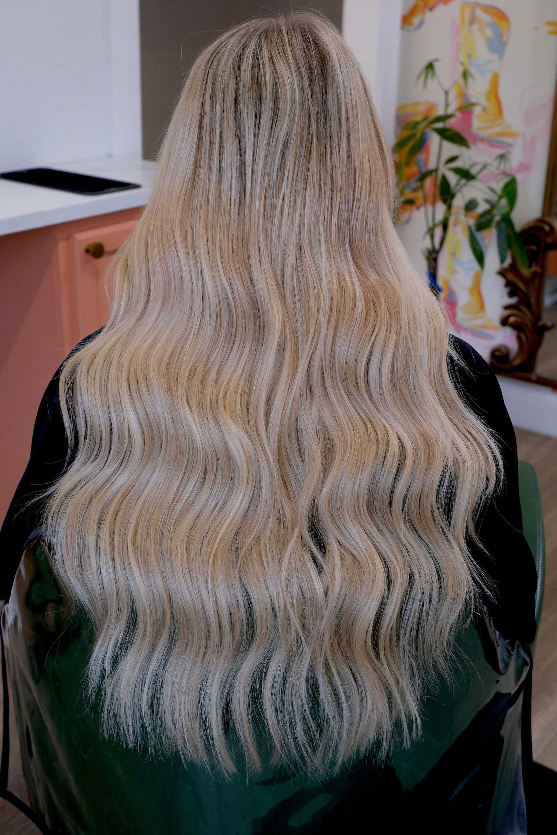Long wavy blonde hair viewed from the back in a salon setting with colorful decor.