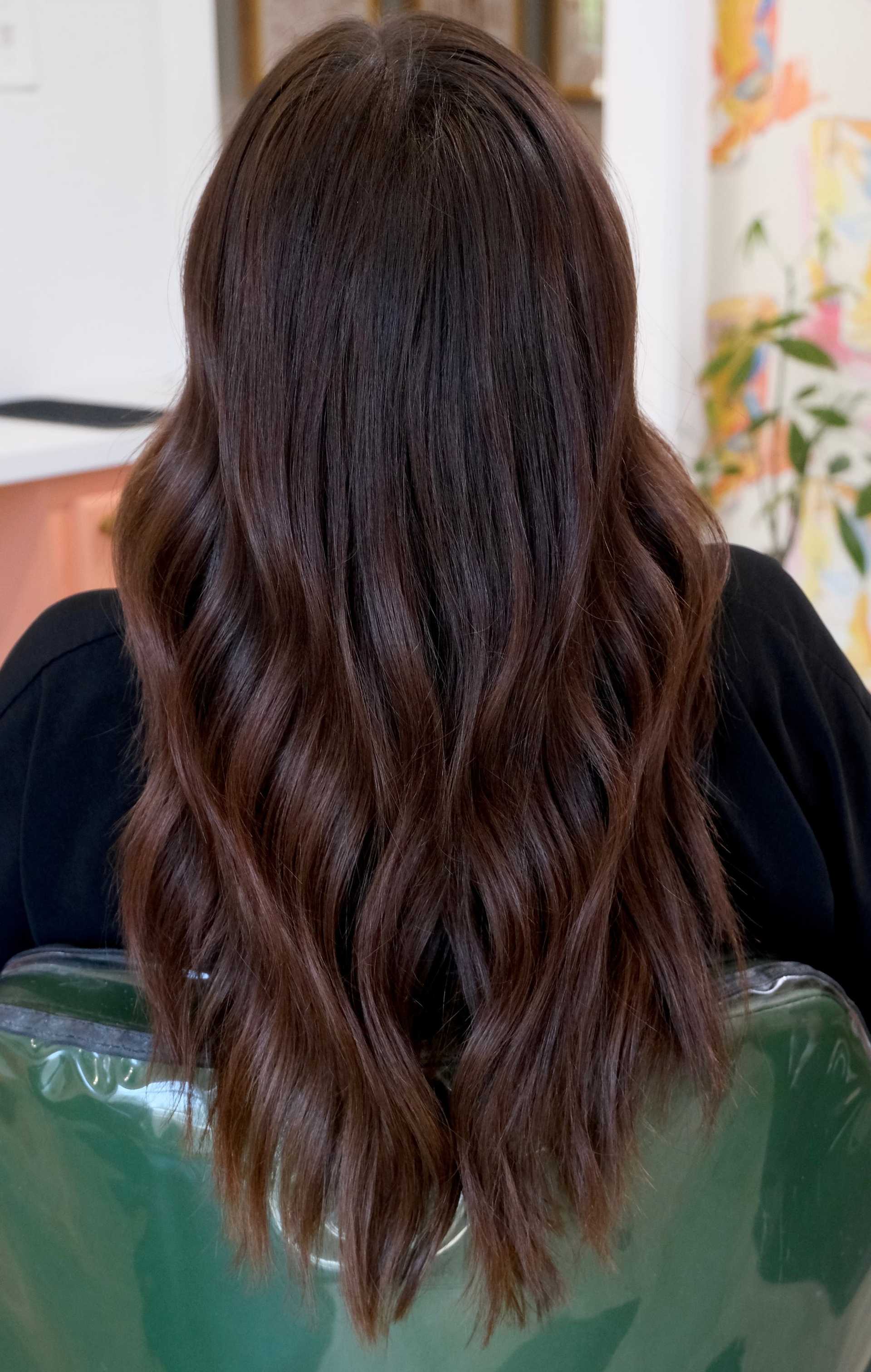 Back view of a person with long, wavy brown hair sitting in a chair indoors.